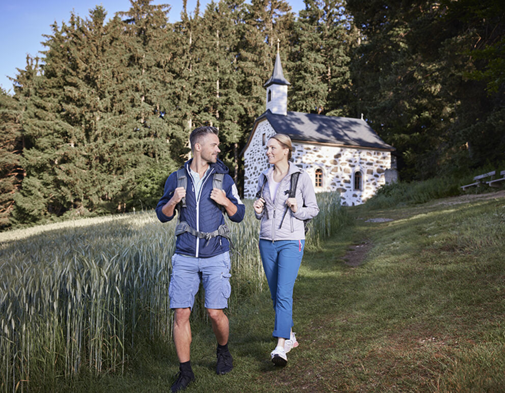 Eine Frau und ein Mann wandern auf einer Wiese. Im Hinergrund siehe man das Kammerer Kreuz, eine Kapelle im Steinblostil erbaut.